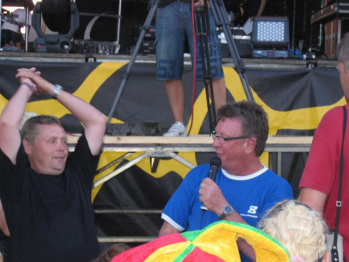 A man in a blue shirt speaks into a microphone on an outdoor stage while another man stands nearby with his arms raised.