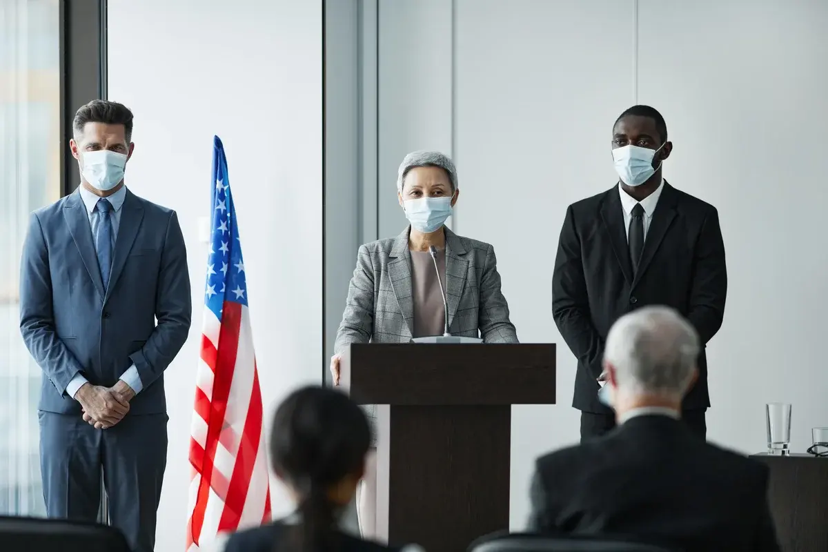 Three masked business leaders stand at a podium beside an American flag during a conference.