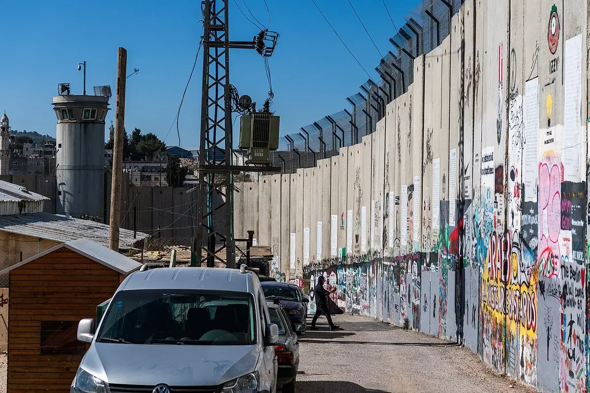 A tall concrete separation wall covered in graffiti runs through Bethlehem beside a narrow street and parked cars.