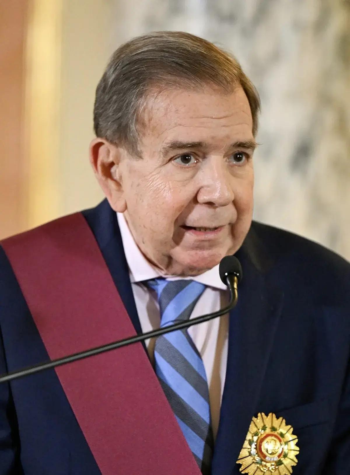 Elderly man in suit with medal and sash speaking at microphone