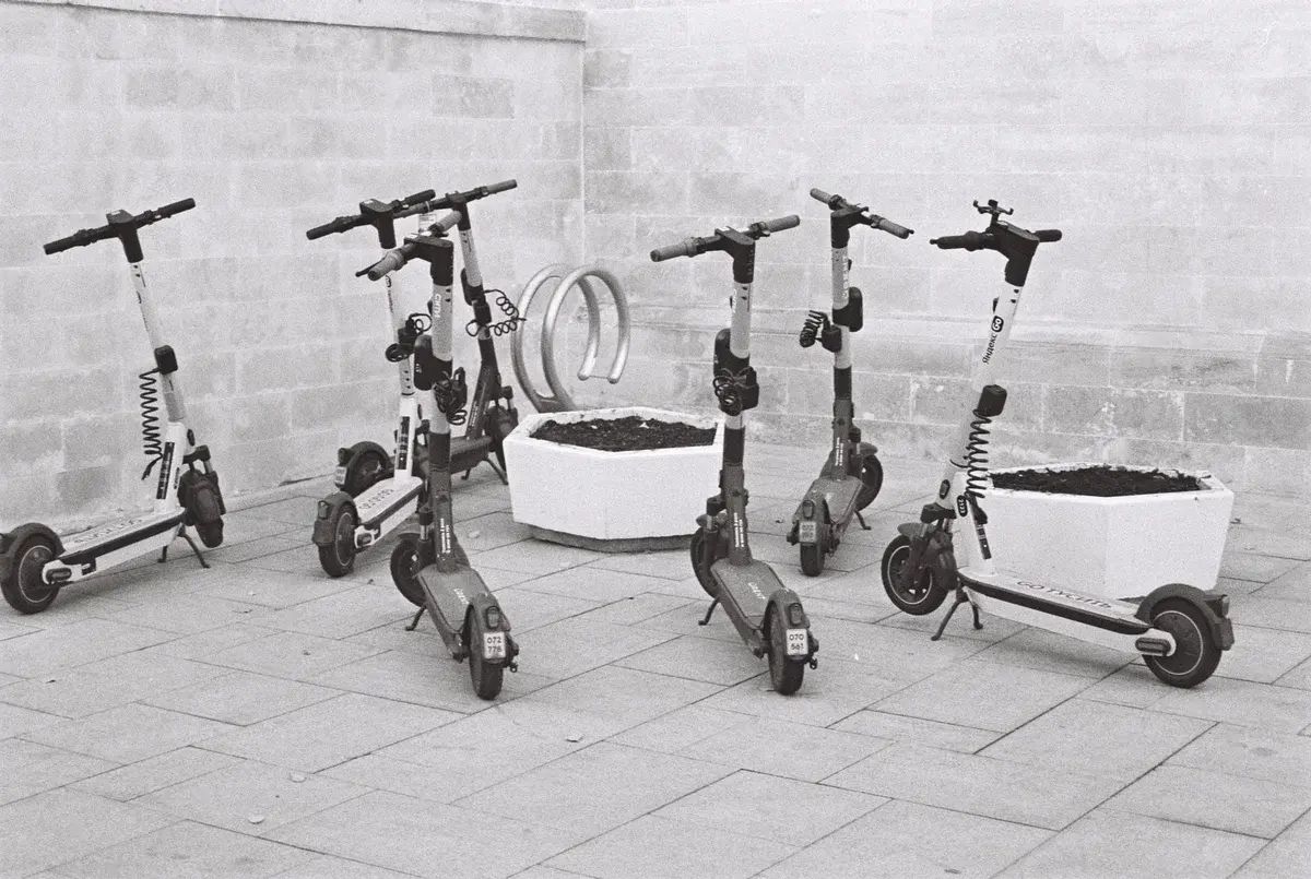 Black and white image of several parked electric scooters beside planters on a tiled pavement.