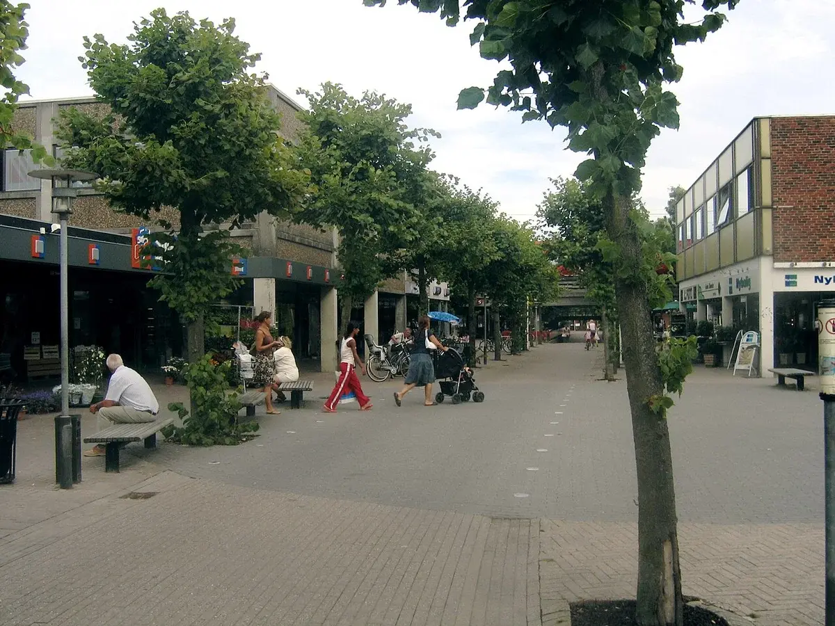 Pedestrian shopping street with trees, benches, shops, and people walking.