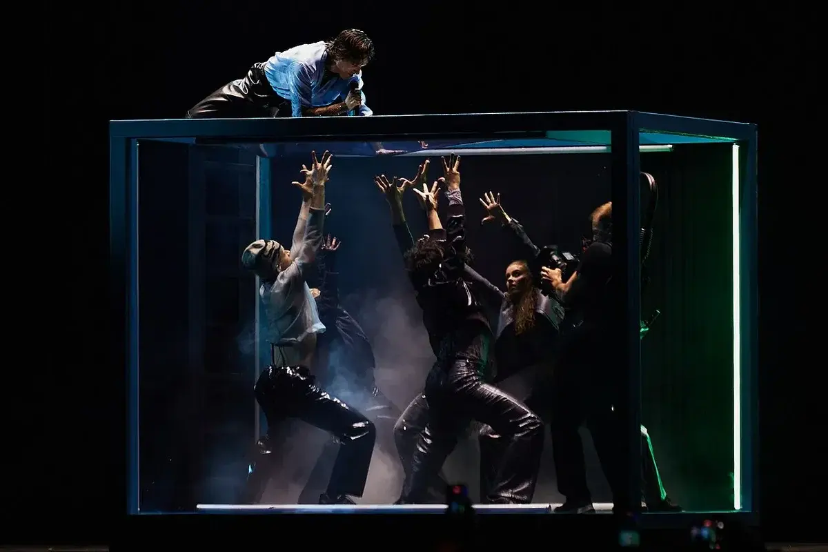 Singer on top of glass box with dancers inside reaching up
