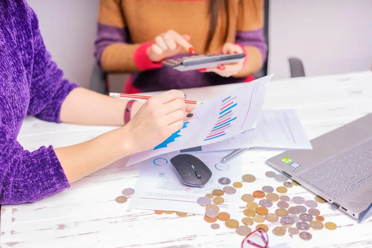 Two women reviewing financial charts at an office desk with a laptop and coins.