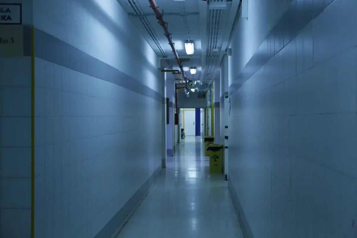 An empty, dimly lit hospital corridor with tiled walls and ceiling lights.