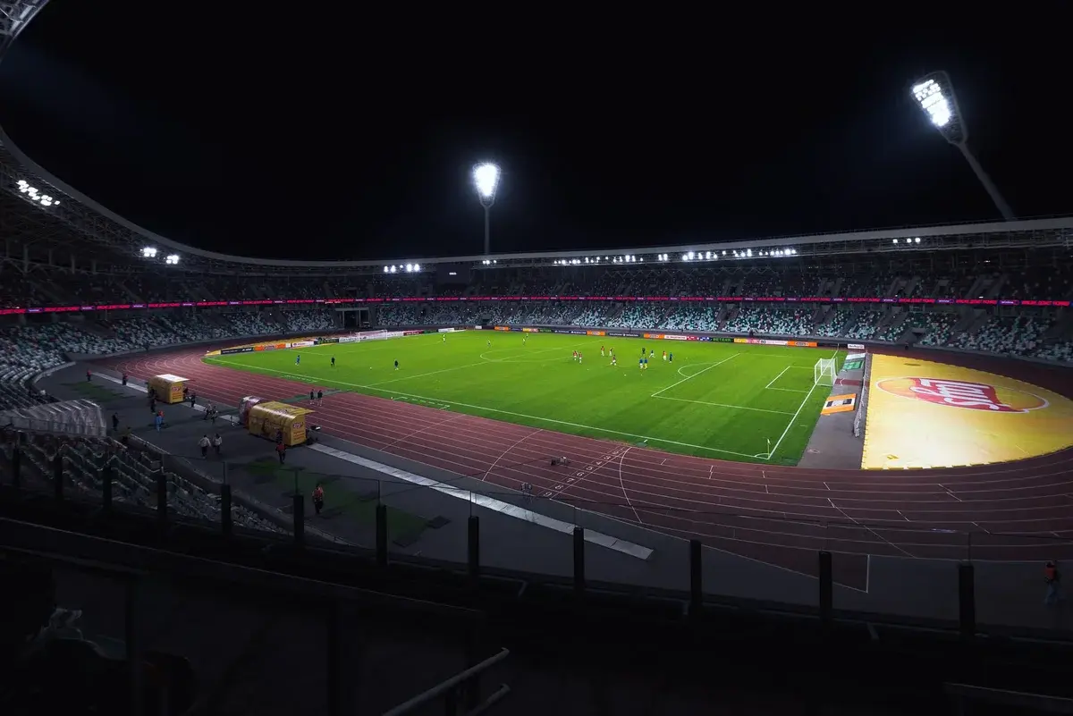 A night view of a football stadium with players on the field and empty stands.