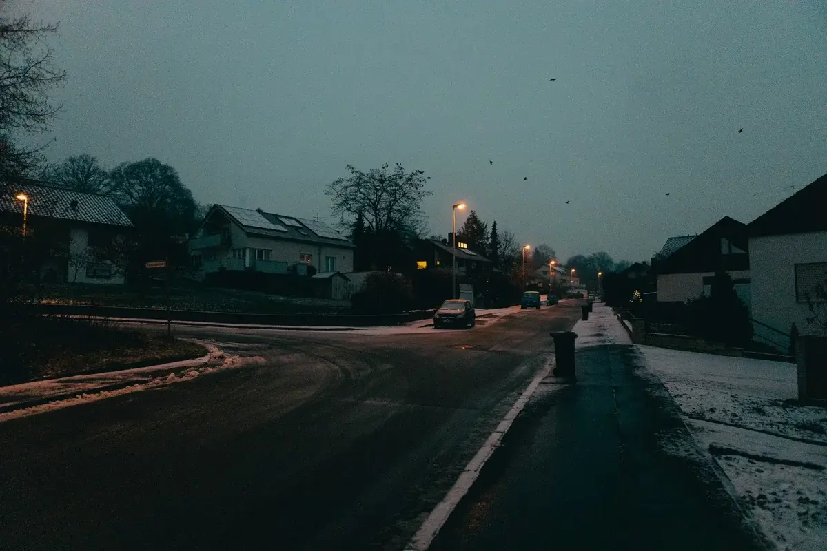 A snowy suburban street at dusk with streetlights and houses.