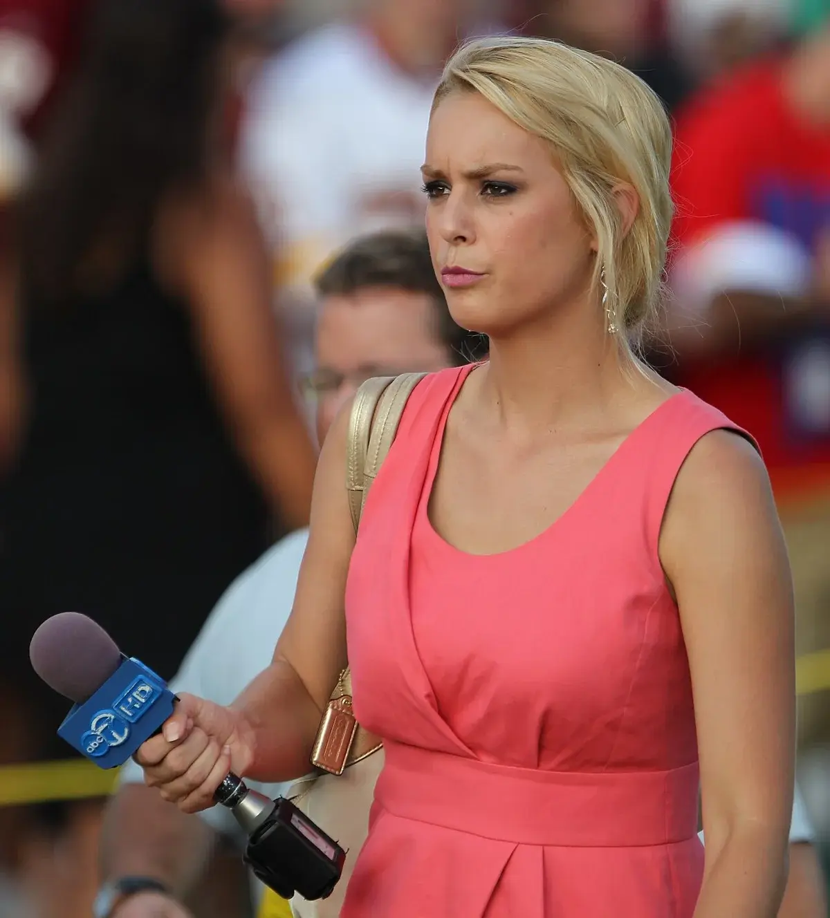A blonde sports reporter holding a microphone at a football training camp.