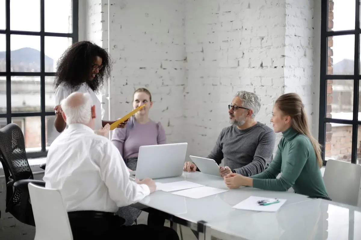 A Black woman presents to coworkers seated around a table in a bright office.