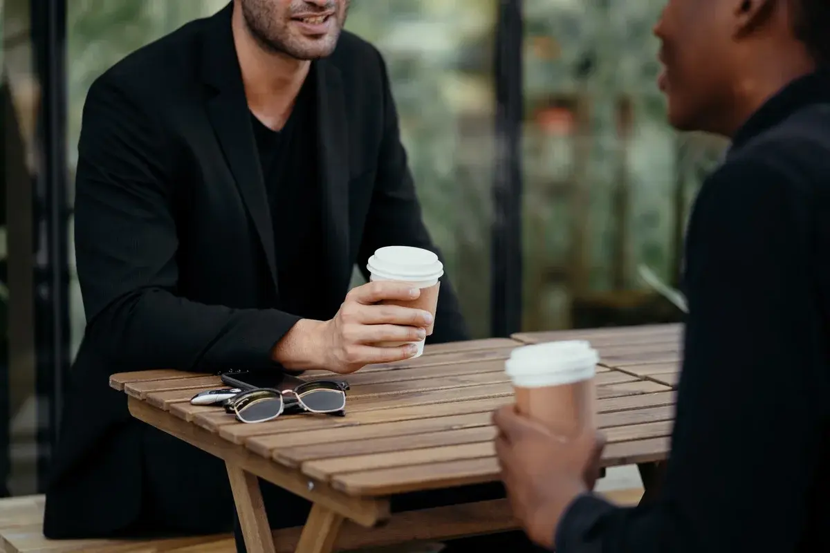 Two adults holding takeaway coffee cups at an outdoor table.