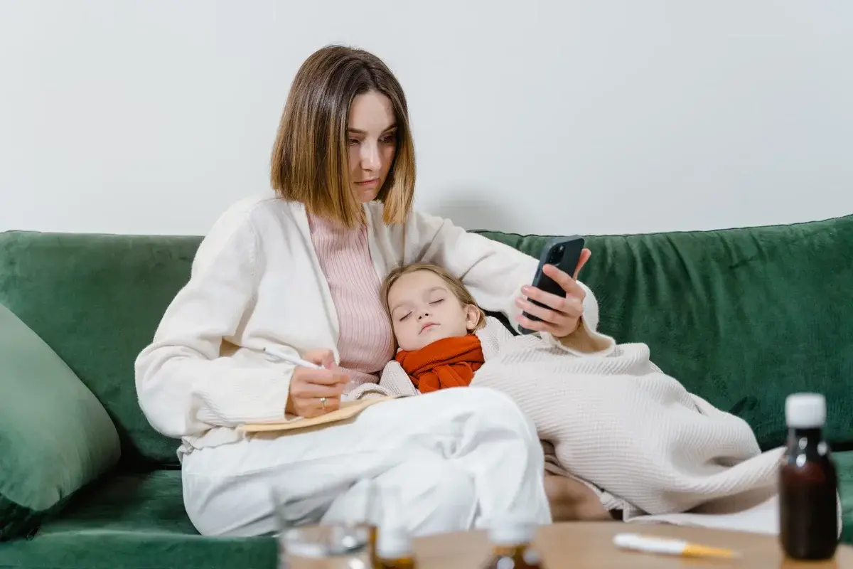 A worried mother sits beside a sleeping child on a sofa while checking symptoms on her phone.