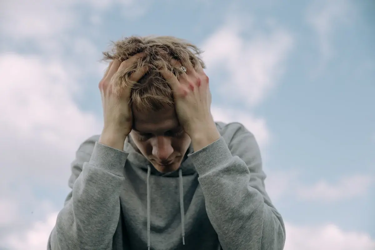 A young man in a gray hoodie holds his head against a cloudy sky.