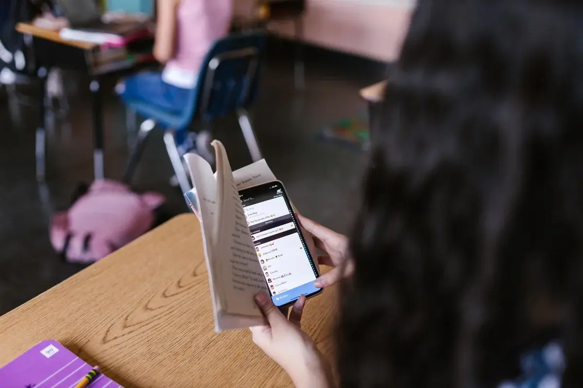 A student in a classroom reads a book while looking at a smartphone.